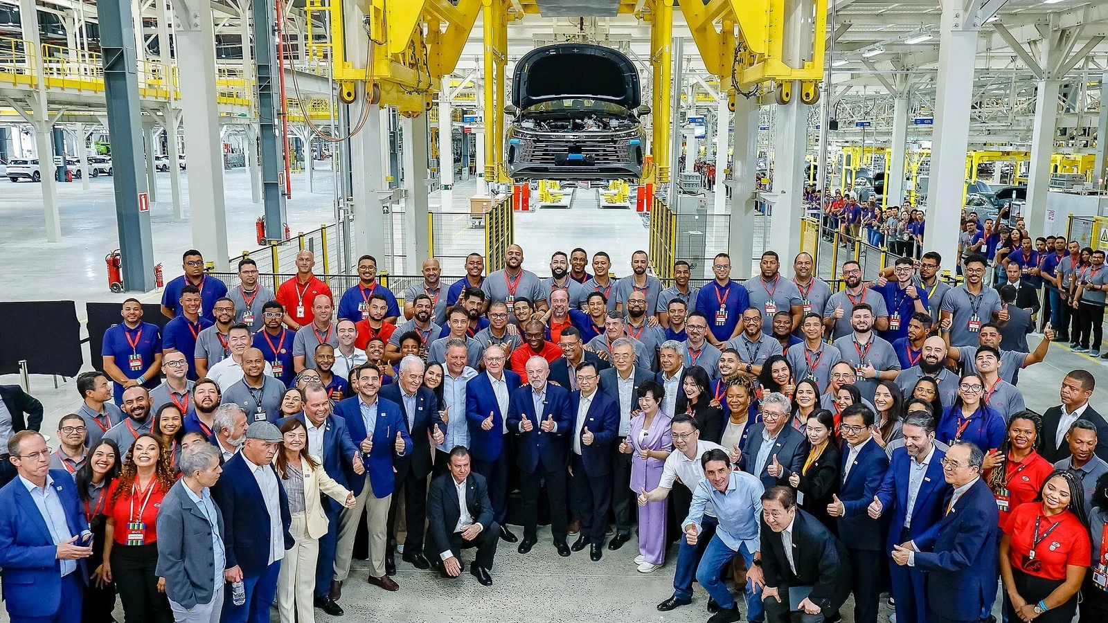 Funcionários e executivos reunidos na linha de montagem da fábrica da BYD em Camaçari, na Bahia, com um carro suspenso ao fundo
