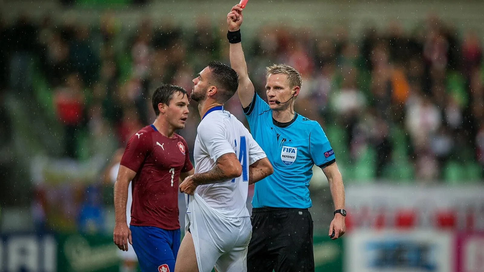 Árbitro de camisa azul mostra o cartão vermelho a um jogador durante partida de futebol sob chuva