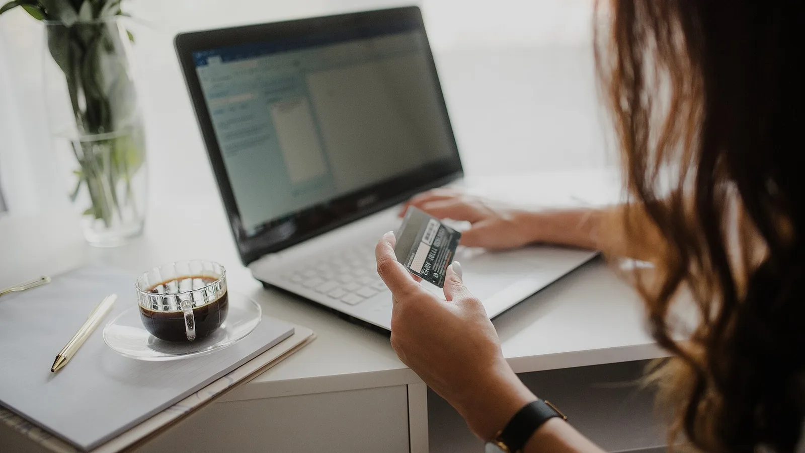 Mulher segura um cartão de crédito enquanto usa um notebook sobre uma mesa branca