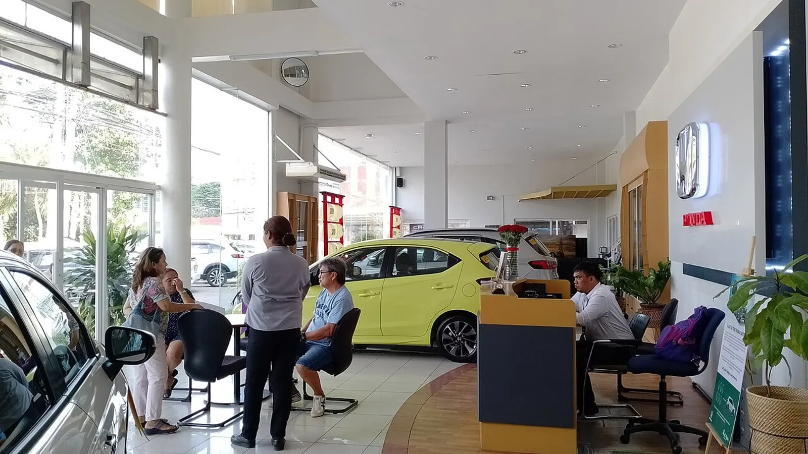 Salão de uma concessionária com um hatch amarelo exposto e clientes sendo atendidos