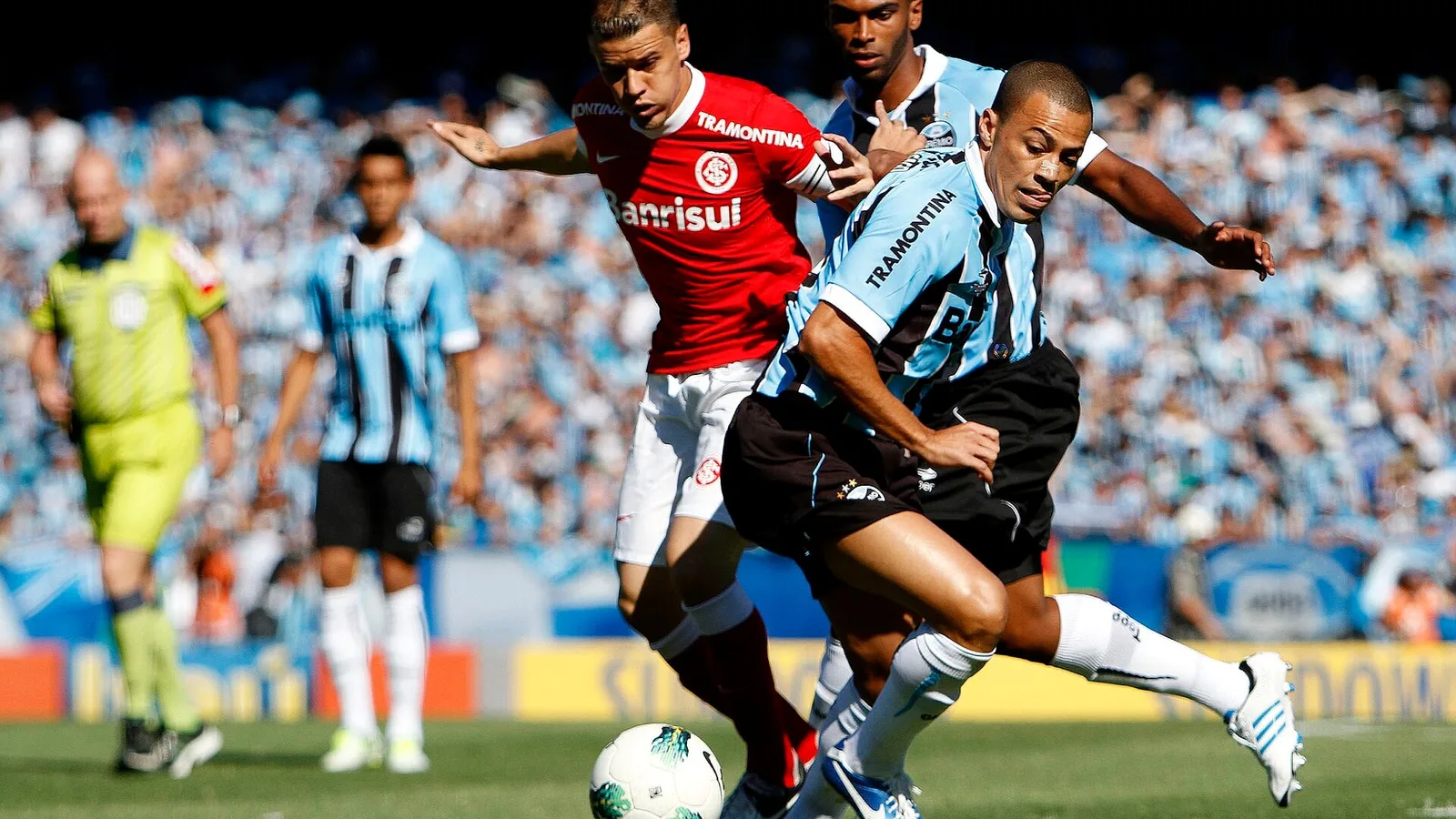 Jogadores de Internacional e Grêmio disputam a bola diante da arquibancada lotada, com o árbitro ao fundo