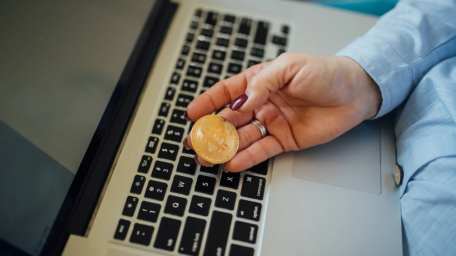Mão segurando uma moeda física dourada com o símbolo do bitcoin sobre o teclado de um notebook