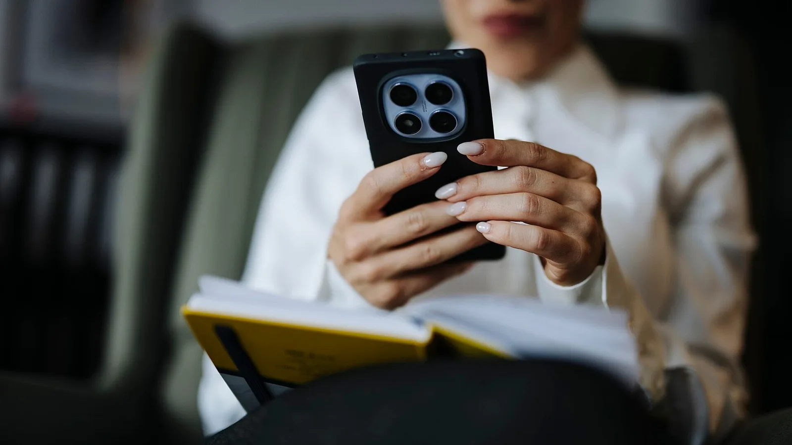 Mulher segurando um celular com as duas mãos enquanto lê, sentada, com um caderno no colo