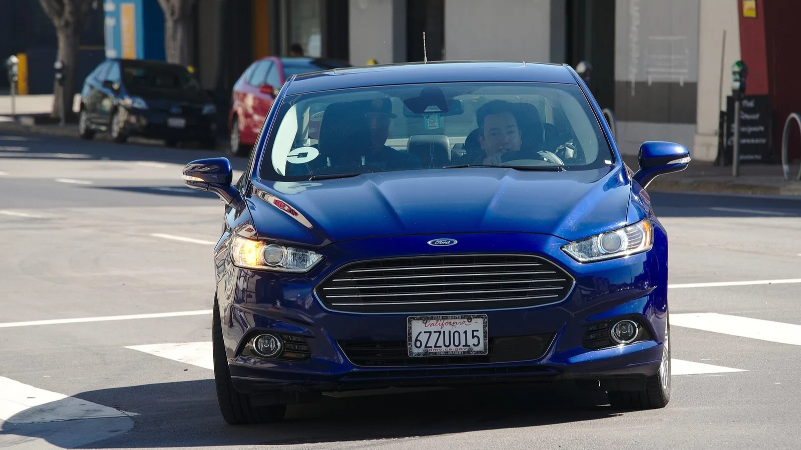 Sedã Ford Fusion azul com adesivo de aplicativo de transporte no para-brisa, visto de frente em uma rua