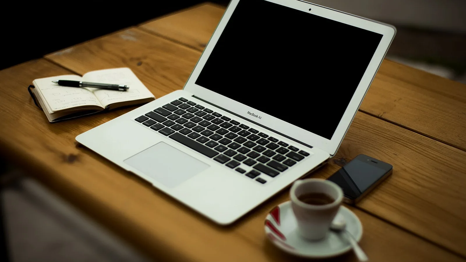 Notebook aberto sobre uma mesa de madeira, com caderno, caneta, celular e uma xícara de café