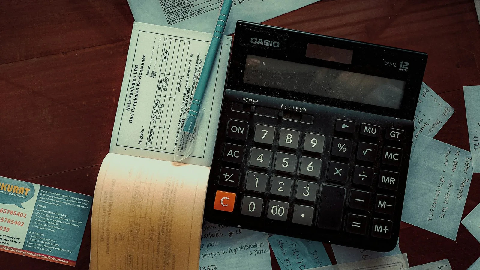 Calculadora sobre uma mesa de madeira, com caneta, bloco de notas e vários recibos e papéis com anotações de contas