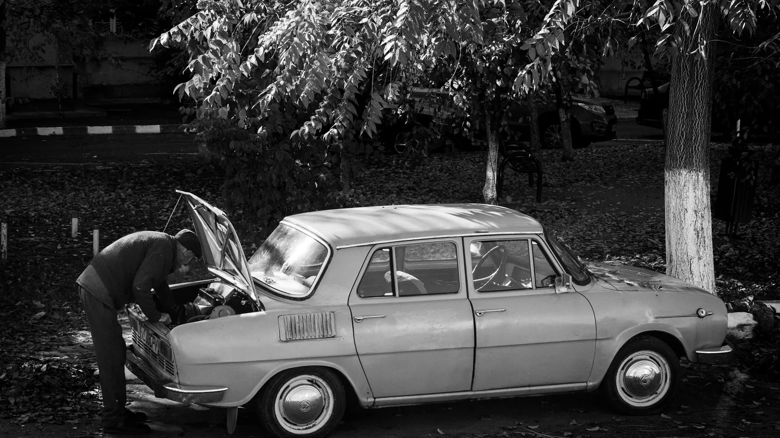 Homem inclinado sobre o motor de um carro antigo estacionado, com o capô aberto, em foto em preto e branco