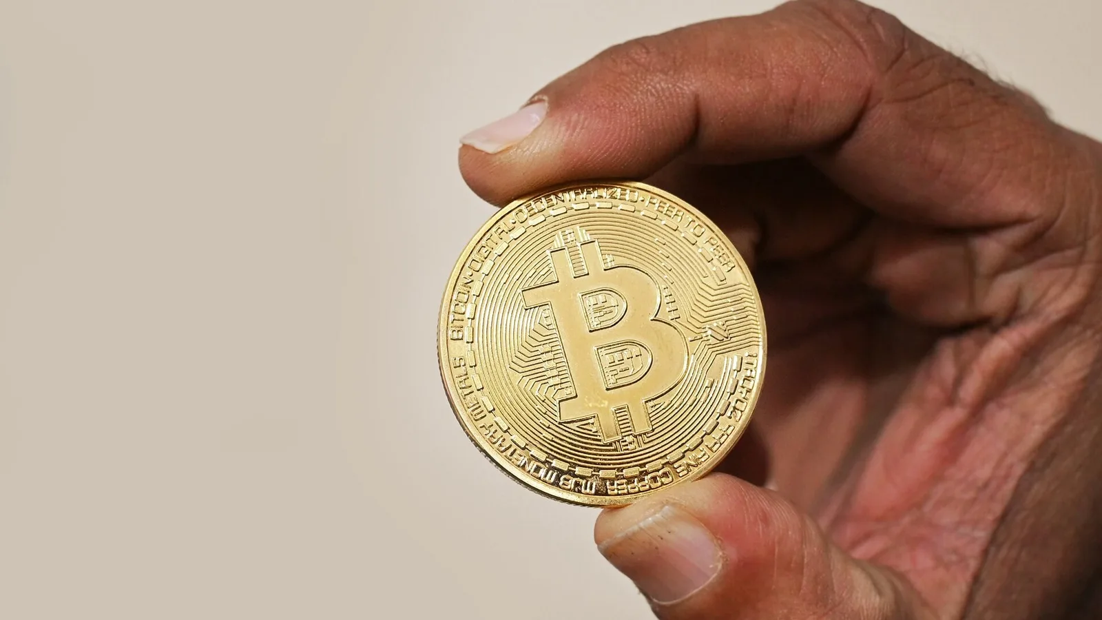 Mão segurando uma moeda física dourada com o símbolo do bitcoin, usada como ilustração de criptoativos